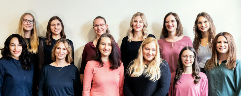 Zwölf Frauen stehen in zwei Reihen vor einer hellen Wand und lächeln in die Kamera. Sie tragen legere, bunte Oberteile und haben unterschiedliche Frisuren, was eine fröhliche und freundliche Gruppenatmosphäre schafft. - Logopädische Praxis Christina Kopp