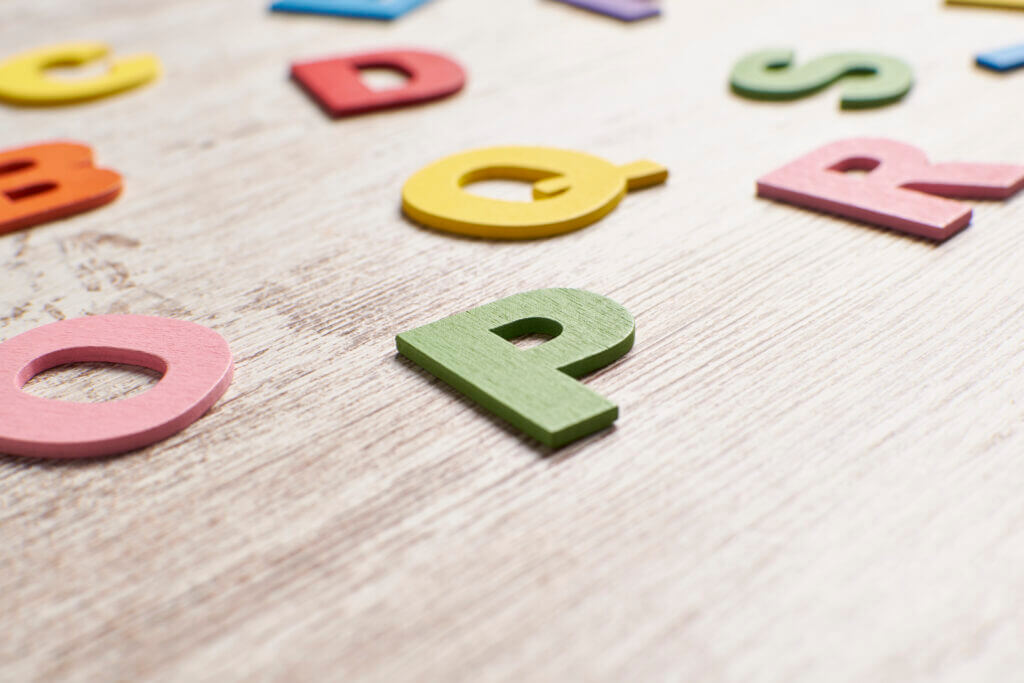 Bunte Holzbuchstaben, verstreut auf einer hellen Holzfläche, wobei der grüne Buchstabe P im Fokus steht und die anderen Buchstaben im Hintergrund verschwommen sind. - Logopädische Praxis Christina Kopp