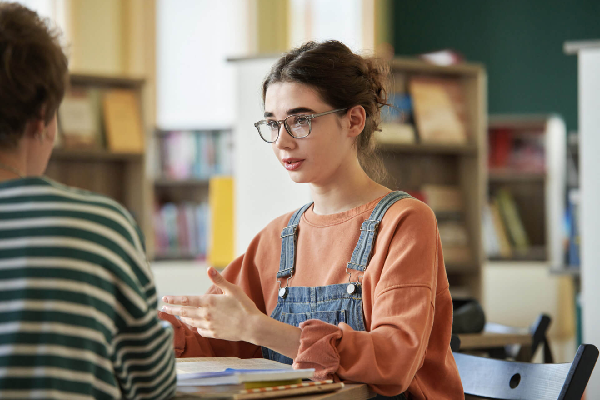 Eine junge Frau mit Brille und Latzhose sitzt an einem Tisch in einer Bibliothek und unterhält sich, wobei sie mit den Händen gestikuliert, mit einer anderen Person, die der Kamera den Rücken zuwendet. Im Hintergrund sind Bücherregale zu sehen. - Logopädische Praxis Christina Kopp