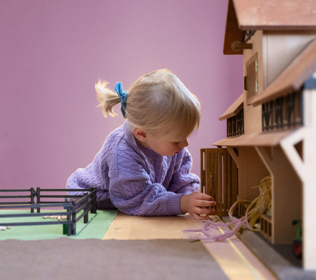 Ein kleines Kind mit blonden Haaren und einer blauen Schleife spielt auf einem Tisch mit einem Spielzeugstall aus Holz; es trägt einen lila Pullover und steht vor einer rosa Wand. - Logopädische Praxis Christina Kopp