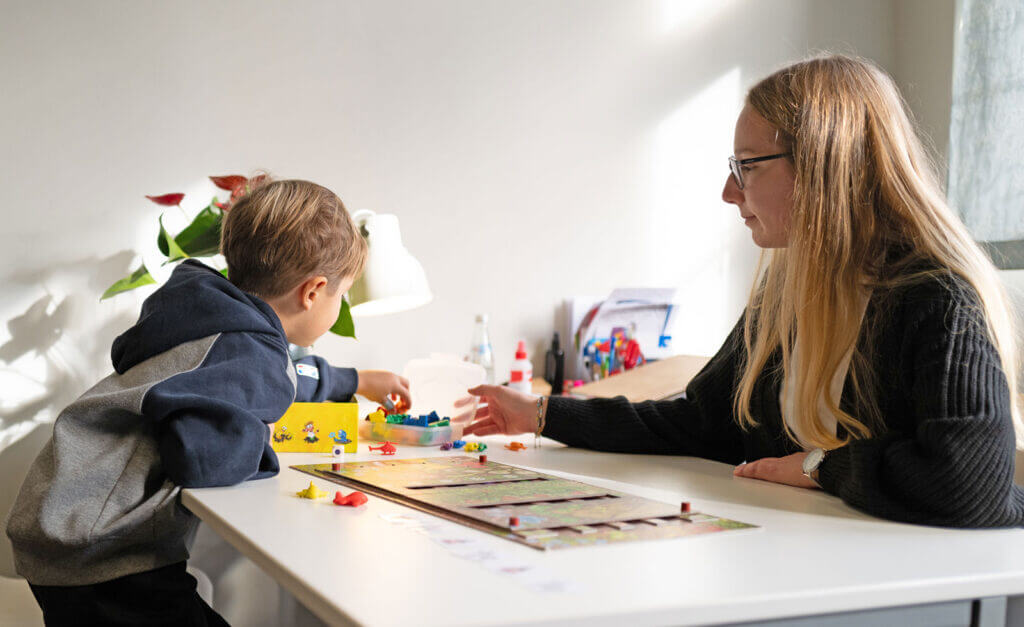 Eine Frau und ein kleiner Junge sitzen an einem weißen Tisch und spielen gemeinsam ein Brettspiel. Sonnenlicht fällt herein, und im Hintergrund sind Malutensilien zu sehen, was eine warme Atmosphäre schafft, die ideal ist, um die Kommunikation zu fördern – perfekt für alle, die auf der Suche nach einem Logopädie-Anbieter in Weiden sind. - Logopädische Praxis Christina Kopp