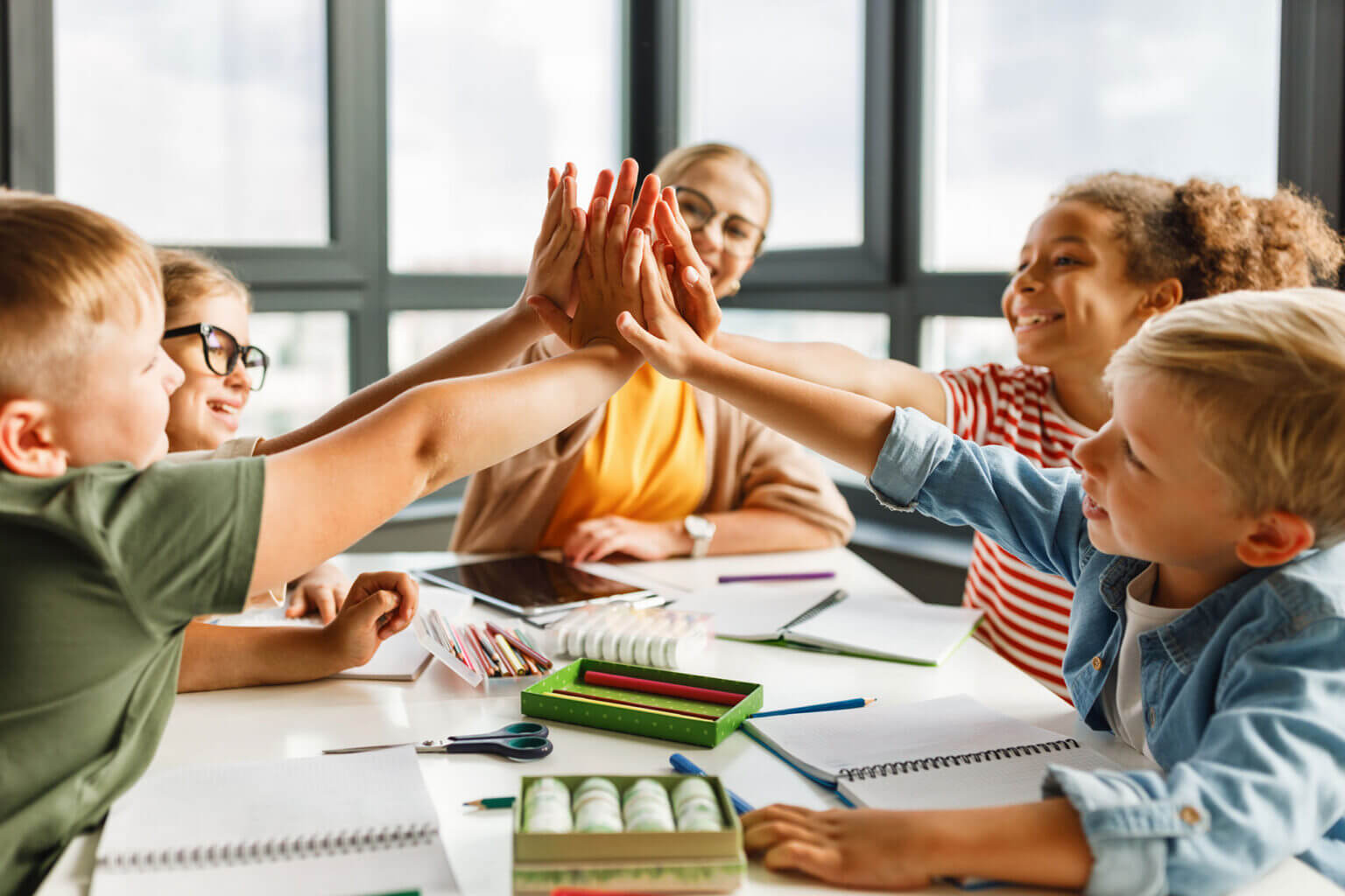 Vier lächelnde Kinder sitzen um einen Tisch herum und klatschen sich gegenseitig ab, während im Hintergrund ein Erwachsener zuschaut und lächelt. Auf dem Tisch liegen Schulmaterialien und Hefte verstreut, was eine lebhafte Atmosphäre schafft, die sich perfekt für Inhouse-Seminare in der Kita oder Schule eignet. - Logopädische Praxis Christina Kopp