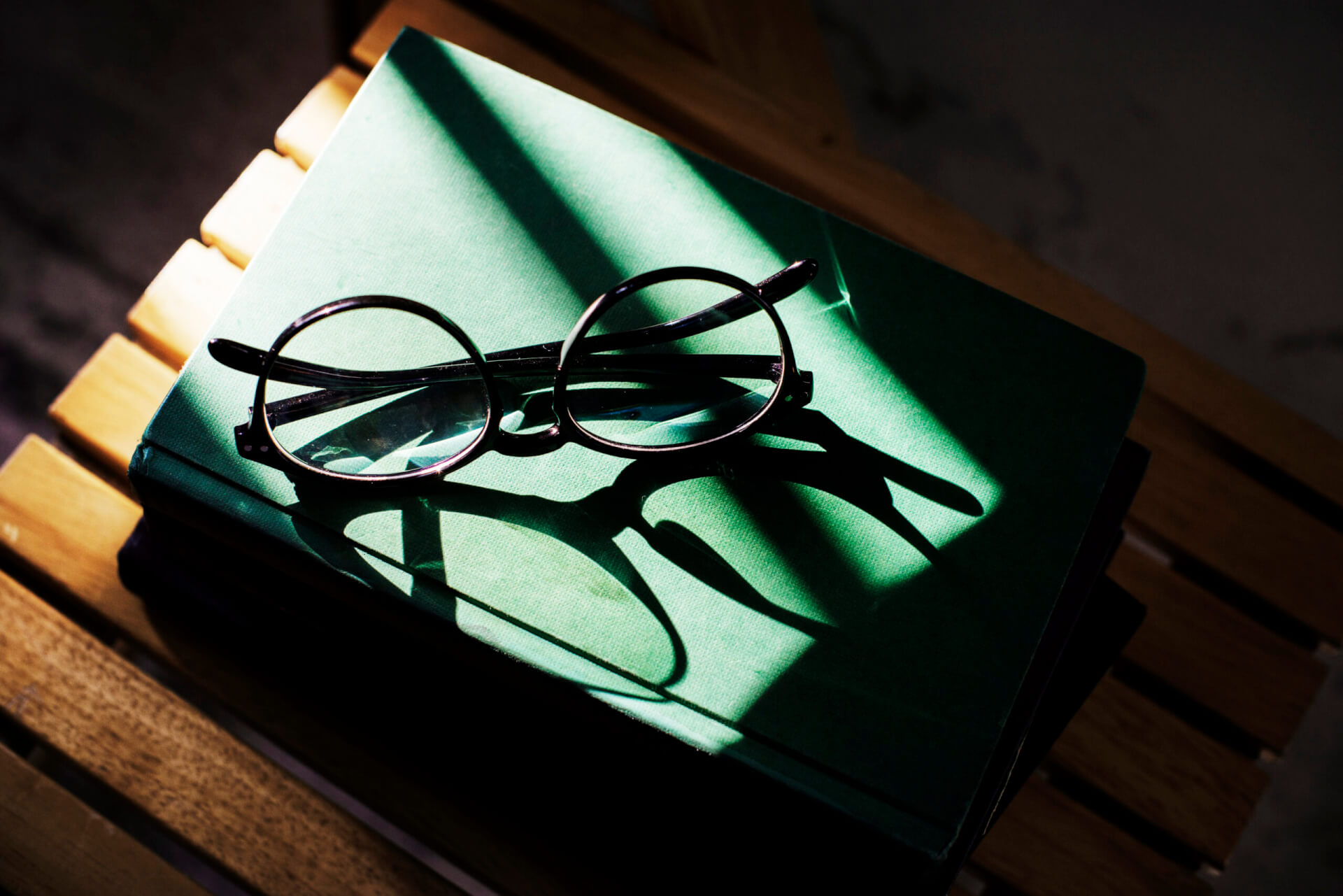 Eine runde Brille liegt auf einem geschlossenen grünen Buch, das auf einer Holzfläche steht. Ein Sonnenstrahl wirft Schatten auf das Buch und die Brille und schafft so eine ruhige, besinnliche Stimmung. - Logopädische Praxis Christina Kopp