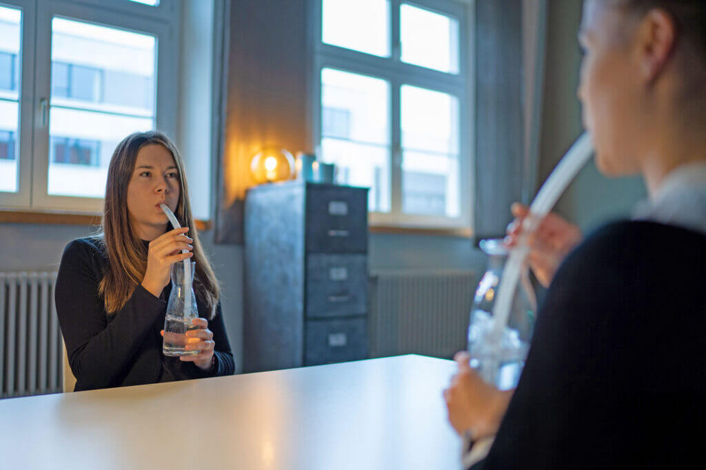Zwei Personen sitzen sich an einem Tisch gegenüber und verwenden während einer Sitzung zur Logopädie bei Stimmstörungen Inhalationsgeräte aus Glas. - Logopädische Praxis Christina Kopp