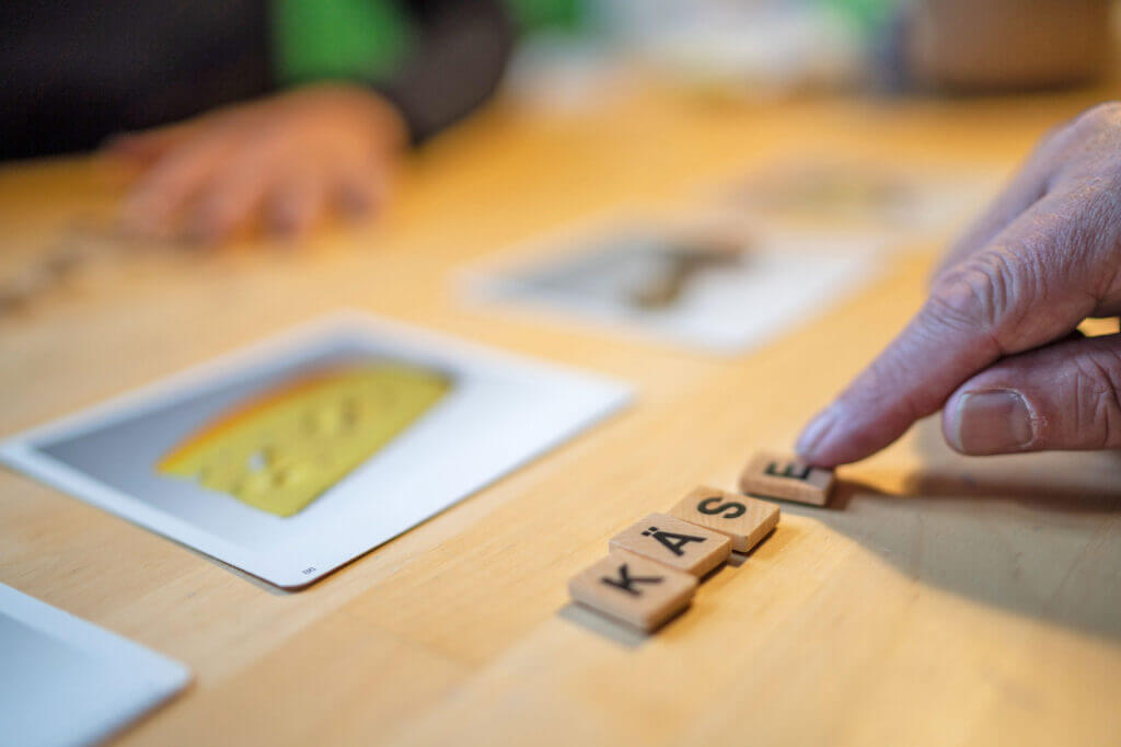 Eine Hand legt die letzte Buchstabenkachel, um das Wort KÄSE zu bilden, auf einen Tisch neben eine Bildkarte mit Käse – eine gängige Übung in der neurologischen Logopädie. - Logopädische Praxis Christina Kopp