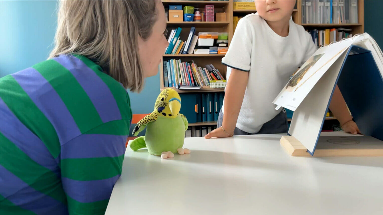 Eine Frau und ein kleiner Junge schauen sich während einer Sitzung zur Logopädie bei Kommunikationsstörungen ein Buch auf einem Ständer an, während zwischen ihnen auf einem Tisch ein Plüschsittich sitzt. - Logopädische Praxis Christina Kopp