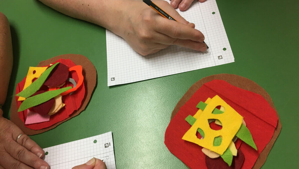 Hände, die auf Millimeterpapier zeichnen, neben zwei Bastelsandwiches aus Filz auf einem grünen Tisch – als Symbol für die Arbeit an Störungen des Schriftspracherwerbs. - Logopädische Praxis Christina Kopp