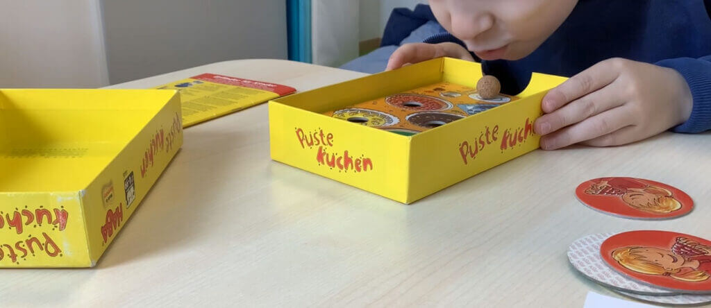 Ein Kind bläst auf eine kleine Korkkugel in der gelben Schachtel des Brettspiels Puste Kuchen – ein Hilfsmittel, das häufig zur Behandlung von Sprachstörungen bei Kindern eingesetzt wird. - Logopädische Praxis Christina Kopp
