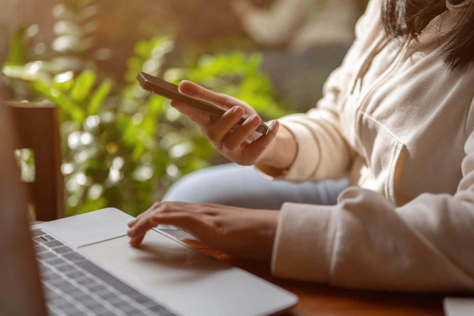 Eine Person, die einen hellen Kapuzenpulli trägt, benutzt mit einer Hand ein Smartphone, während sie mit der anderen auf einem Laptop tippt; sie sitzt an einem Fenster, im Hintergrund sind Grünpflanzen zu sehen. - Logopädische Praxis Christina Kopp